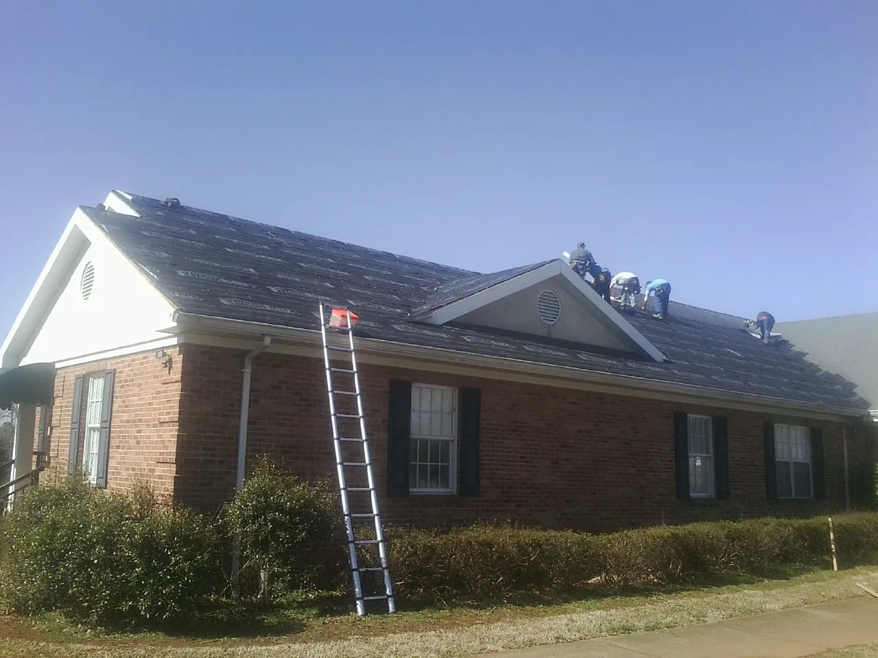 Crew tearing shingles off a brick ranch roof with a ladder set — Metal Roof Repair work by our crew in Leon